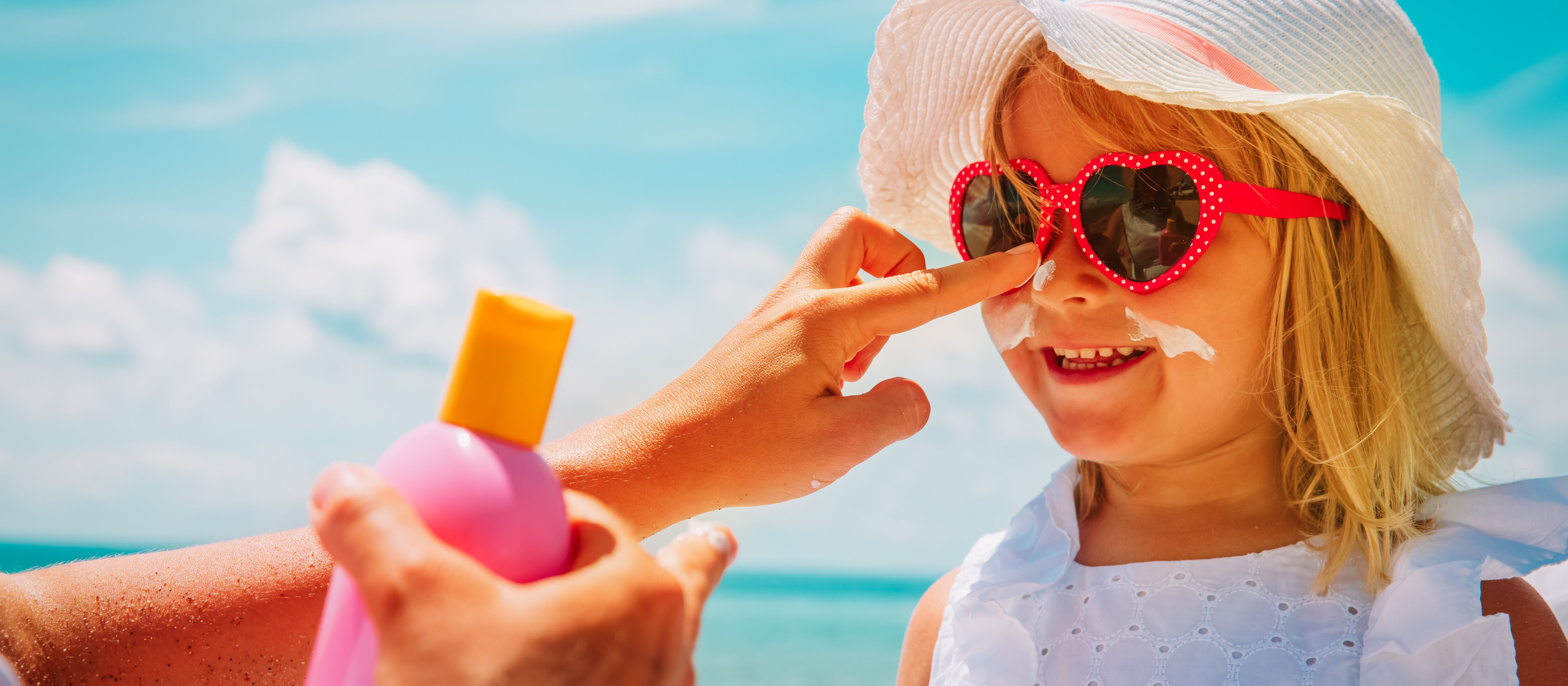 Blonde girl wearing white dress and sunhat, pink sunglasses, getting sunscreen applied to her face