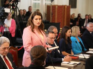 Woman with brown here and pink blazer standing up and speaking at a board table, surrounded by other people, media cameras in the background