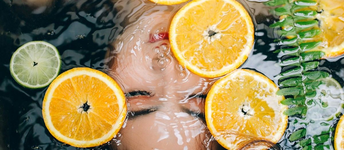 Woman with dark hair is submerged in bath water with lemons and herbs floating on the water