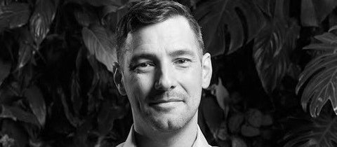 Black and white image shows man with his arms crossed, looking at the camera, with leafy trees in the background