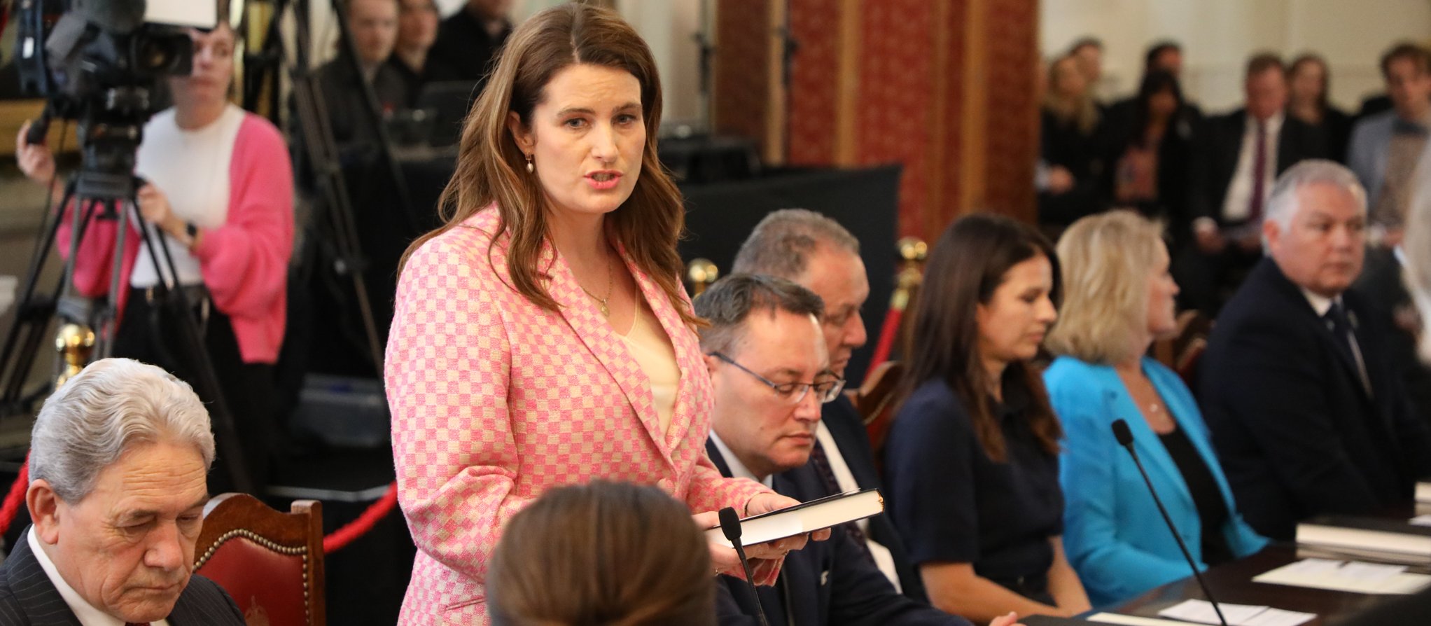 Woman with brown here and pink blazer standing up and speaking at a board table, surrounded by other people, media cameras in the background