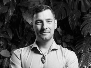 Black and white image shows man with his arms crossed, looking at the camera, with leafy trees in the background