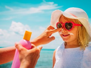 Blonde girl wearing white dress and sunhat, pink sunglasses, getting sunscreen applied to her face